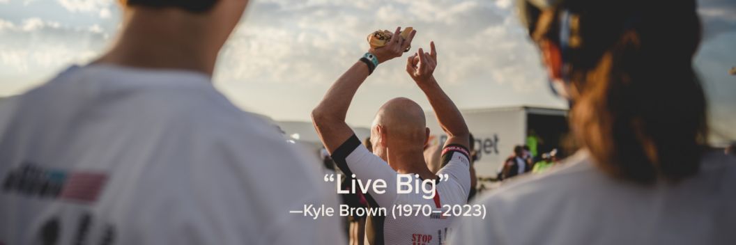 A photo of Kyle Brown raising his arms in success after completing his triathlon. His quote “Live Big” is next to 1970–2023 in parenthesis.